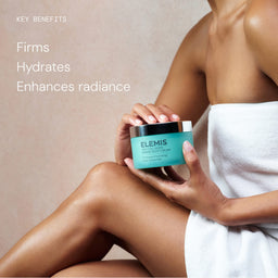 A woman wrapped in a white towel holds an Elemis skincare cream jar against a soft beige background with text reading 'KEY BENEFITS' and 'Firms Hydrates Enhances radiance.'.