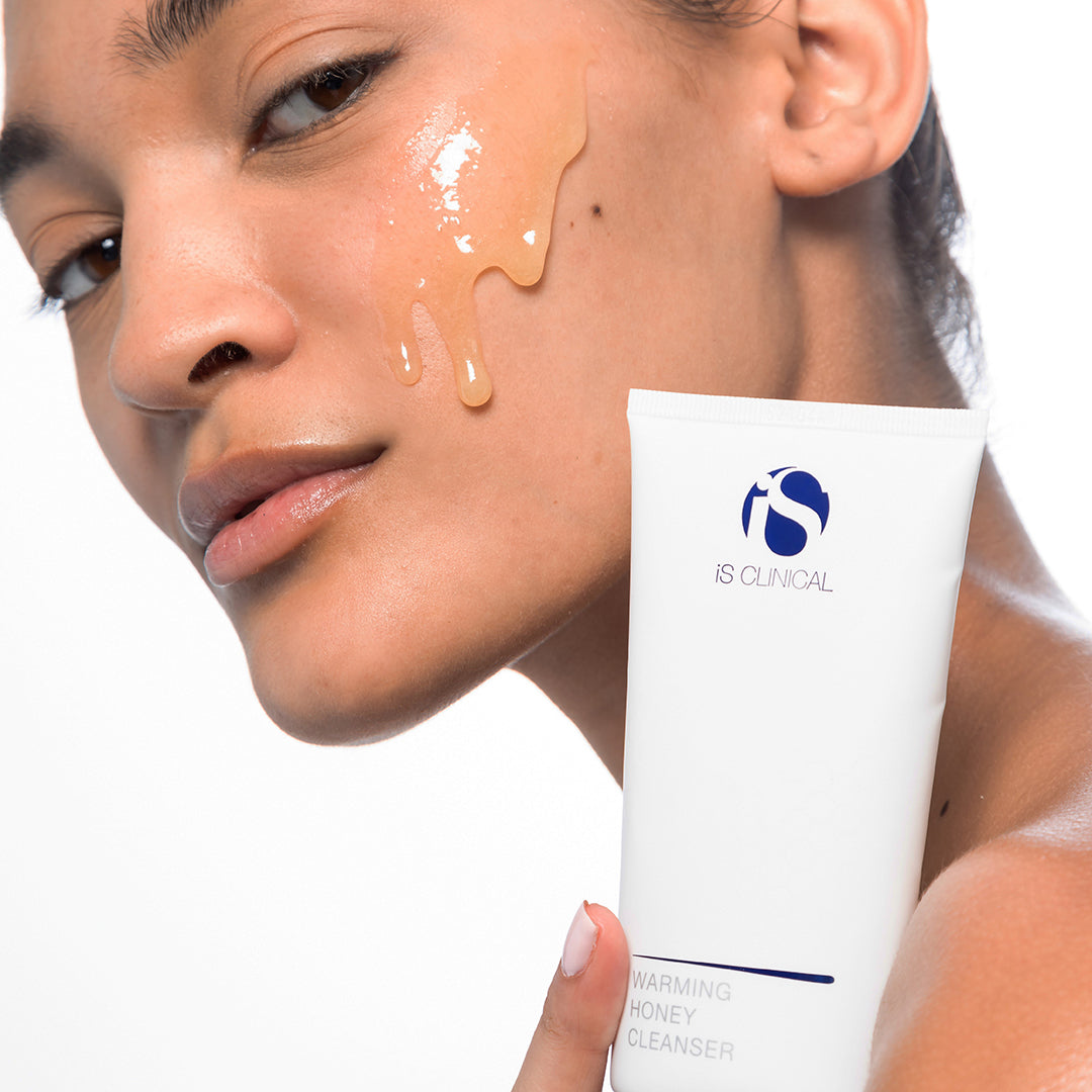 Woman applying a cosmetic product to her face with a tube labeled 'iS Clinical' in the foreground.