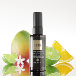 A bottle of ghd hair color developer with a black cap, styled with fresh mango, lime, orange, and a small white flower on a reflective white surface against a light gray background.