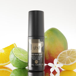 A bottle of ghd hair color, specifically the 'naturally even after' developer, styled with citrus slices, a mango, and a white flower on a reflective white surface against a white background.