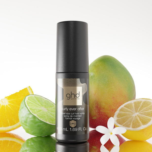 A bottle of ghd hair color, specifically the 'naturally even after' developer, styled with citrus slices, a mango, and a white flower on a reflective white surface against a white background.