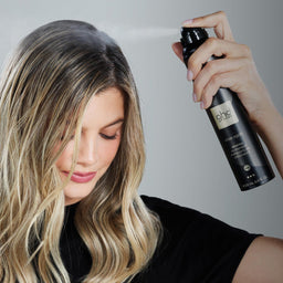 Woman applying hair product spray to her hair against a plain background
