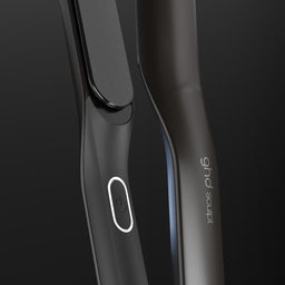 Close-up of a sleek dark gray hair straightener with a glossy black paddle and a white oval logo, set against a dark gradient background. The brand name 'sophie' is visible on the handle.