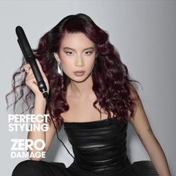 A woman with long, wavy burgundy hair holds a black hair straightener in a studio setting. She is wearing a black strapless top, and the text 'PERFECT STYLING ZERO DAMAGE' is visible on the left side against a gray gradient background.