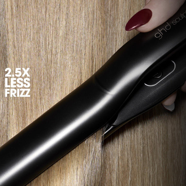 A close-up of a black ghd hair straightener held by a hand with dark red nail polish, resting on a light brown wood grain surface. White text on the left reads '2.