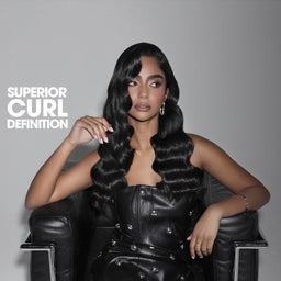 A woman seated in a black chair wearing a voluminous, long, black, defined curling hairstyle. The text 'SUPERIOR CURL DEFINITION' is printed in white on the left side against a dark gray gradient background.