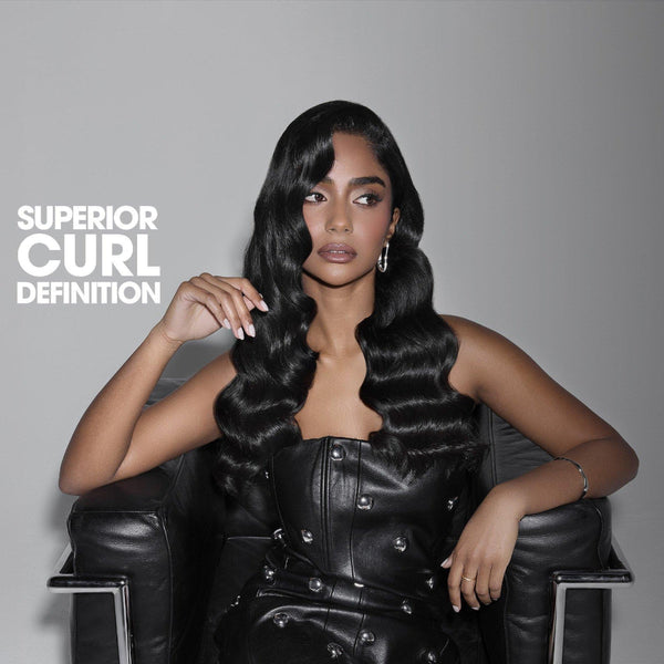A woman seated in a black chair wearing a voluminous, long, black, defined curling hairstyle. The text 'SUPERIOR CURL DEFINITION' is printed in white on the left side against a dark gray gradient background.