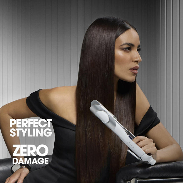 A woman with long, straight dark brown hair sits in a salon-style chair, holding a white hair straightener. She wears a black off-the-shoulder top against a gray paneled background.