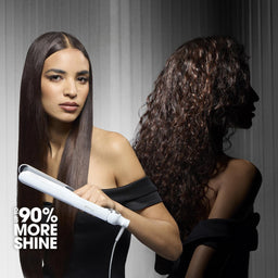 A woman with straight dark hair holds a white hair straightener in front of another woman with long, curled dark hair. The background is a gray vertical-striped studio wall, and the text '90% MORE SHINE' appears in the lower left corner.