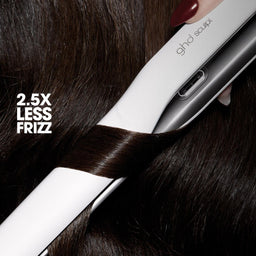 A close-up of a white and silver ghd straightener being used on dark brown hair, with the text '2.5X LESS FRIZZ' overlaid on the left.