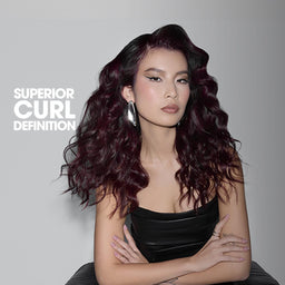 A woman with long, dark, voluminous curled hair wearing a black strapless top, posed against a gray studio background with the text 'SUPERIOR CURL DEFINITION' on the left.