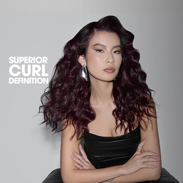 A woman with long, dark, voluminous curled hair wearing a black strapless top, posed against a gray studio background with the text 'SUPERIOR CURL DEFINITION' on the left.