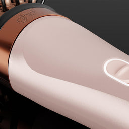 A close-up of a GHD hair styling tool, showing its metallic rose gold barrel, a glowing white power switch, and a copper-colored trim against a dark background.
