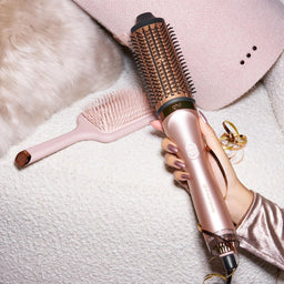 A rose gold hair styling tool with a barrel brush attachment is held by a hand wearing a matching rose gold sleeve, styled on a white textured blanket with a pink hat, a pink brush, and a gold ring nearby.