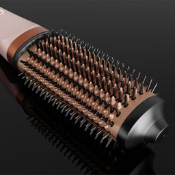A close-up studio shot of a round hairbrush with a pale pink handle, bronze-toned bristles, and a dark metallic end, set against a glossy black background.