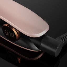 Close-up of a hair straightener with a metallic rose gold body and black accents, shown plugged in on a dark reflective surface.