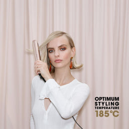 A woman with blonde hair holds a metallic rose gold flat iron against her hair, wearing a white long-sleeve top in front of a light pink curtain backdrop. Text on the right reads 'OPTIMUM STYLING TEMPERATURE 185°C'.