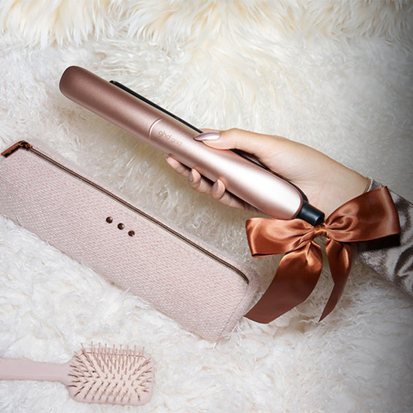 A hand holds a rose gold hair straightener over a white faux fur surface next to a matching light pink travel case and a small hairbrush. The sleeve of the person holding the straightener has a brown satin bow tied around it.