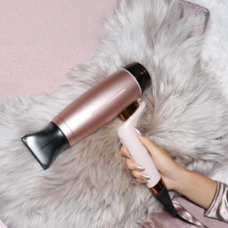 A person's hand holds a pink and black hair styler against a fluffy white faux fur blanket, with a pink satin pillow in the background.
