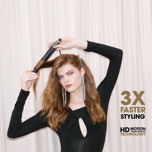 A woman with long wavy light brown hair styling her hair with a black hair straightener, wearing a black long-sleeve top against a cream-colored curtain backdrop. Text overlays on the right read '3X FASTER STYLING' and 'HD MOTION TECHNOLOGY'.