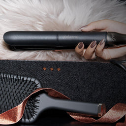 A hand holds a black Ghibliini hair straightener over a fluffy white faux-fur surface, with a black paddle brush and a rose-gold ribbon in the foreground.