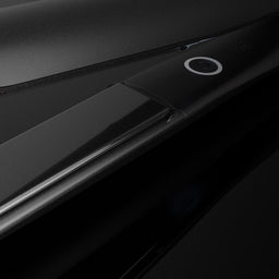 A close-up studio shot of a sleek black hair straightener, showing its smooth metallic plates and a small circular power button on a dark reflective background.