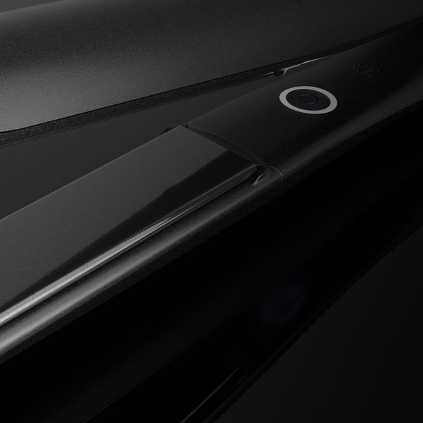 A close-up studio shot of a sleek black hair straightener, showing its smooth metallic plates and a small circular power button on a dark reflective background.
