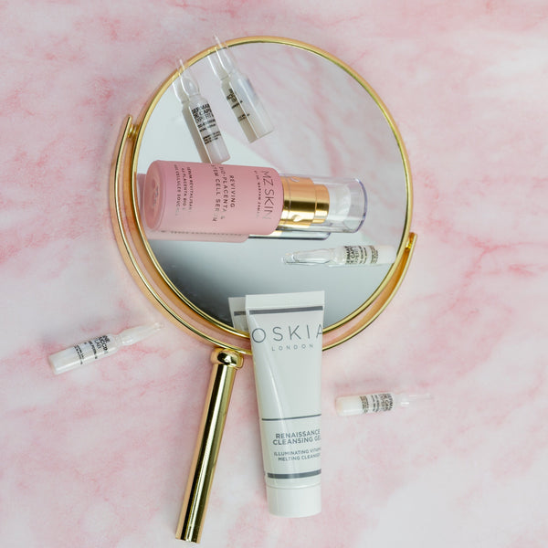 Cosmetic products including a tube of Oskia cream on a pink background with a magnifying mirror.