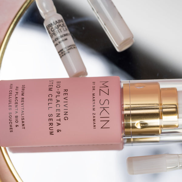 MZ Skin serum bottle with pink cap and gold accents on a reflective surface