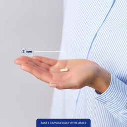 A close-up of a person's hand holding a small white capsule pill, with a measurement overlay indicating 2 mm. The person is wearing a light blue and white striped shirt, and a blue text banner at the bottom reads 'TAKE 1 CAPSULE DAILY WITH MEALS'.