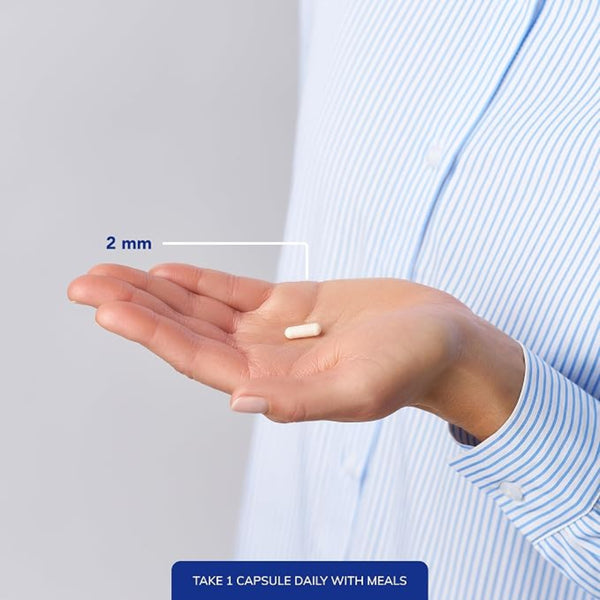 A close-up of a person's hand holding a small white capsule pill, with a measurement overlay indicating 2 mm. The person is wearing a light blue and white striped shirt, and a blue text banner at the bottom reads 'TAKE 1 CAPSULE DAILY WITH MEALS'.