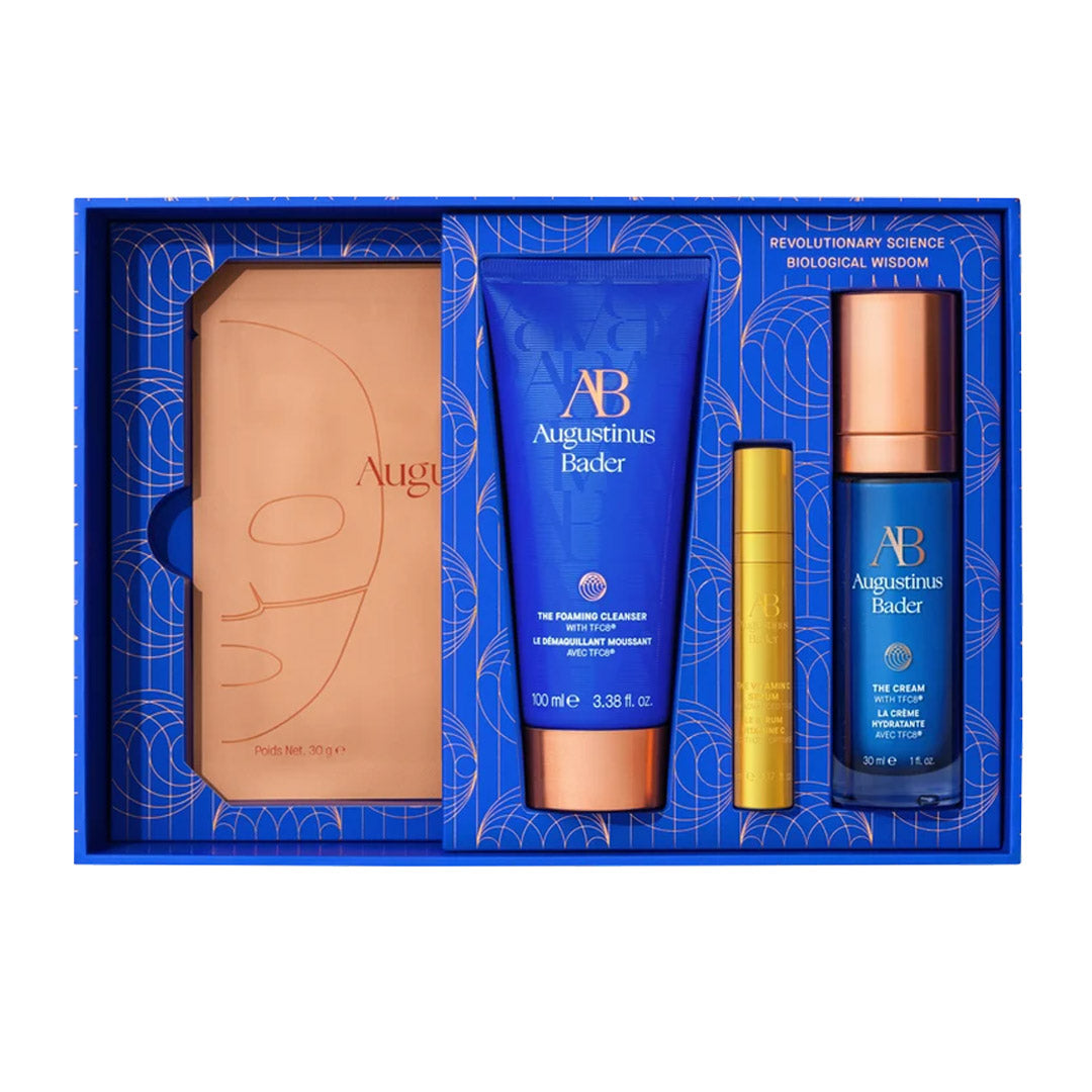 Augustinus Bader gift set in a blue box, featuring a tan leather-like item, a blue cleanser tube, a small gold tube, and a blue bottle with a rose-gold cap.
