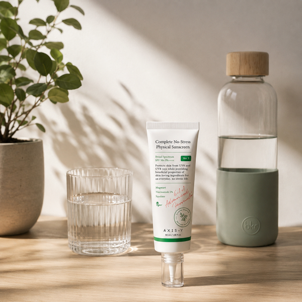 Sunscreen tube, glass water bottle, and glass on a wooden surface with a plant in the background