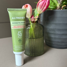 Green skincare tube labeled 'Altruist' next to a green vase with pink flowers on a gray surface.