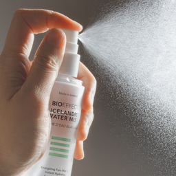 Hand holding a Bioeffect spray bottle with mist being released against a dark background