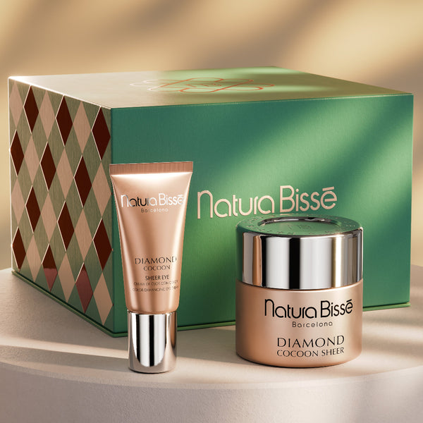 Nature Bisse Diamond Cocoa Shot skincare set featuring a green gift box, a peach-colored cream tube, and a matching jar with a silver lid, displayed on a white pedestal against a warm beige background with soft leaf shadows.