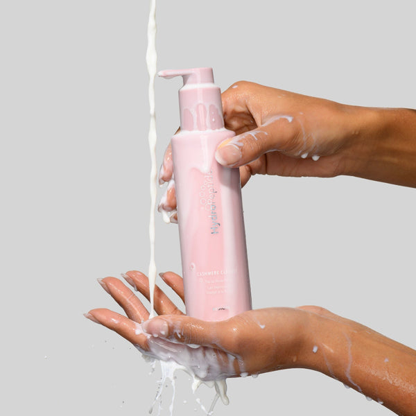 Pink bottle of shampoo being held over a hand with soap suds on a gray background