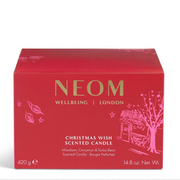 A red rectangular box of NEOM Wellbeing London Christmas Wish Scented Candle, featuring gold and pink lettering and illustrations of a tree and cafe on a white background.