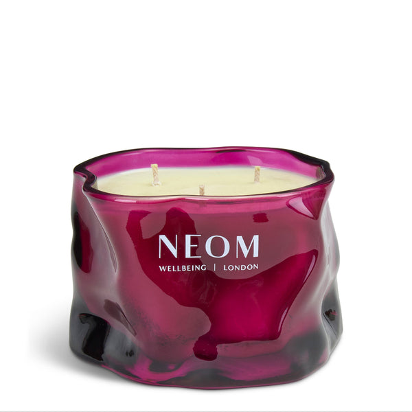 A three-wick scented candle in a wavy, organic-shaped translucent glass jar. The jar has a deep magenta-purple color with a glossy finish and the brand name 'NEOM WELLBEING | LONDON' printed in white on the front.