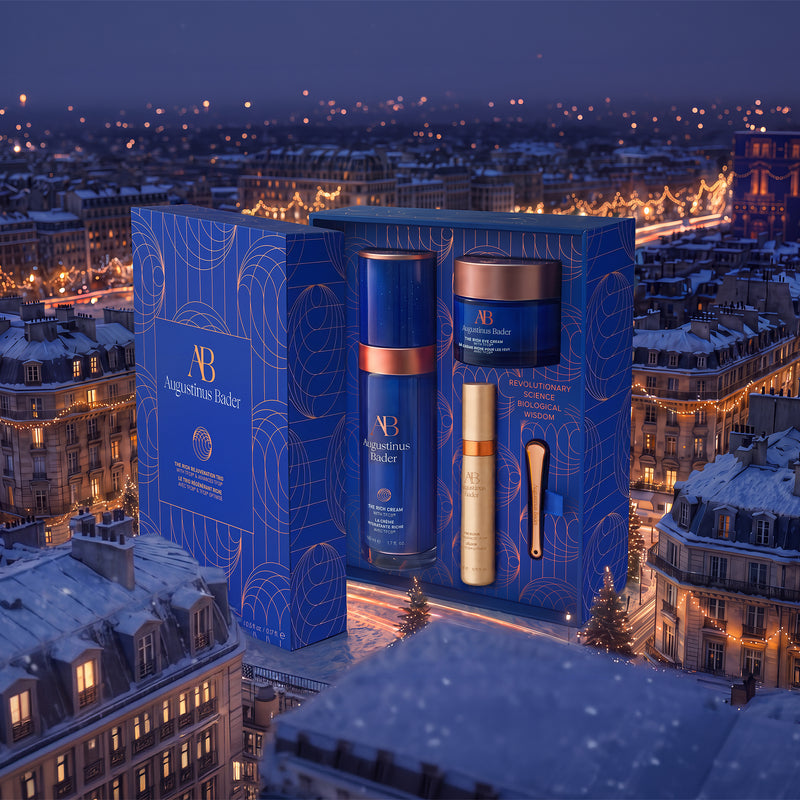 A luxury skincare gift set by Augustinus Bicker, featuring a blue presentation box with gold line art, containing a blue serum bottle with a copper collar, a gold cream bottle, and a gold spatula, set against a Parisian city skyline at night.