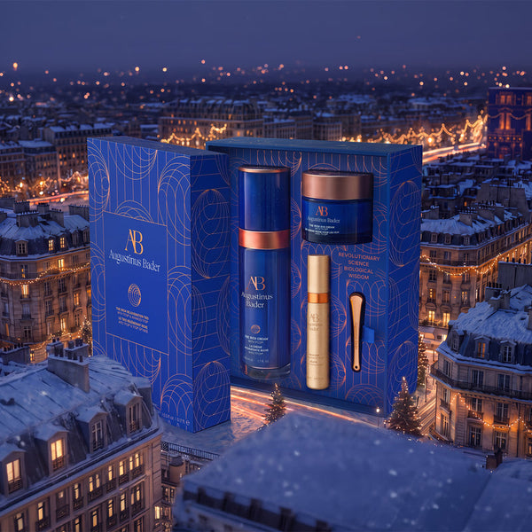 A luxury skincare gift set by Augustinus Bicker, featuring a blue presentation box with gold line art, containing a blue serum bottle with a copper collar, a gold cream bottle, and a gold spatula, set against a Parisian city skyline at night.