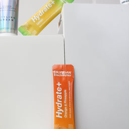 Orange 'Hydrate+' supplement stick pack on a white surface with other supplement packages in the background.