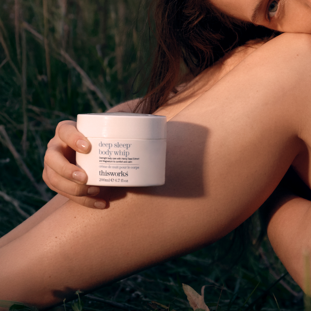 A person outdoors in grass holding a white jar of thisworks Deep Sleep Body Whip, with warm natural sunlight and soft focus on a bare shoulder.