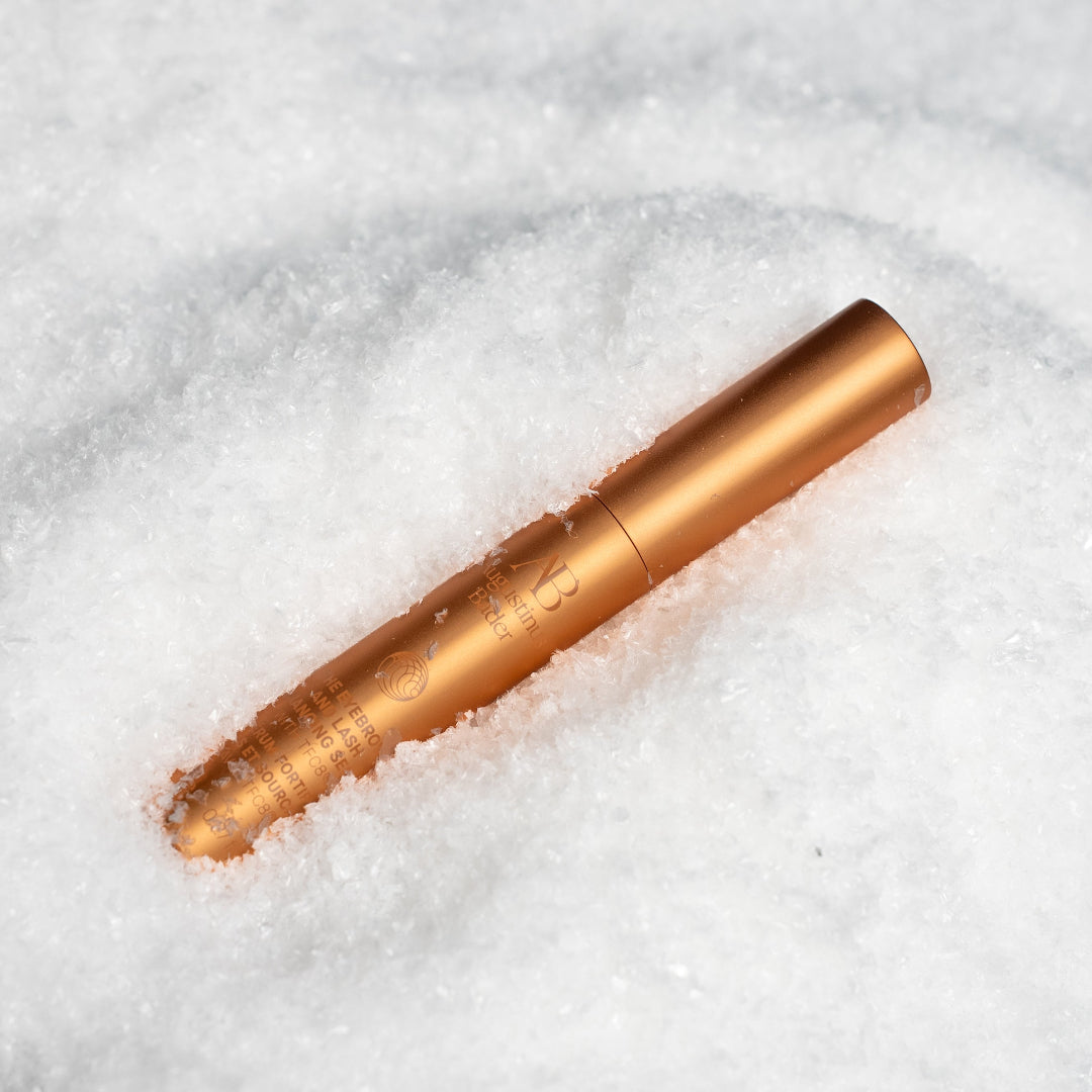 A metallic bronze cosmetic tube lying diagonally in white snow, with embossed text and a circular logo visible on the tube.