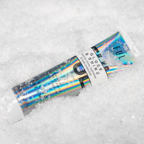 A tube of INFLAME FULL GLAM GLITTER SHINE face makeup powder resting diagonally in white snow. The tube features a vibrant holographic, iridescent finish reflecting blue, green, and pink, with a white paper band label.