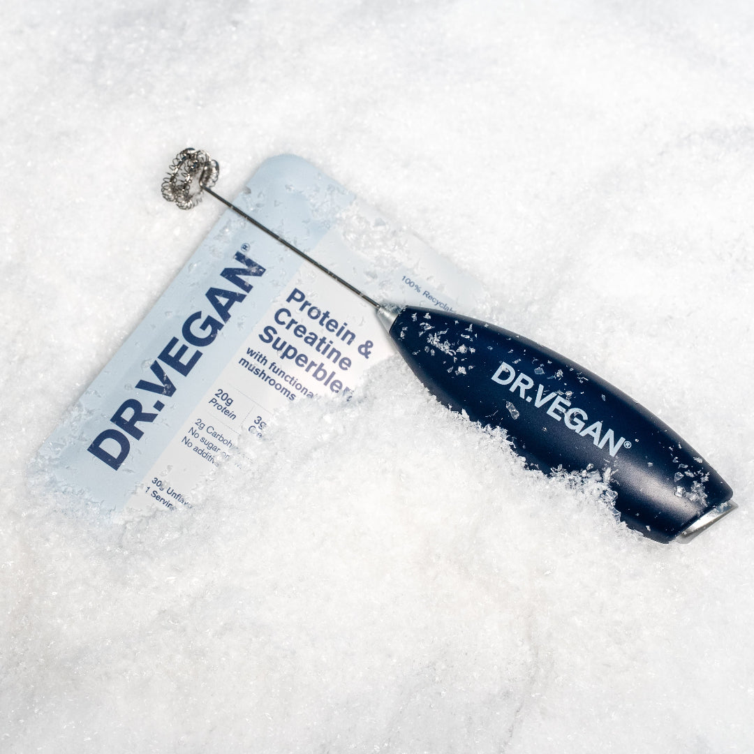 A Dr. Vegan Protein & Creatine Superblend powder pouch lies on white snow with a small metal frother resting on top of the package.