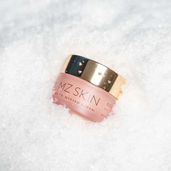 A pink cosmetic jar with a metallic gold lid, labeled 'MZ SKIN', resting on a bed of white snow.