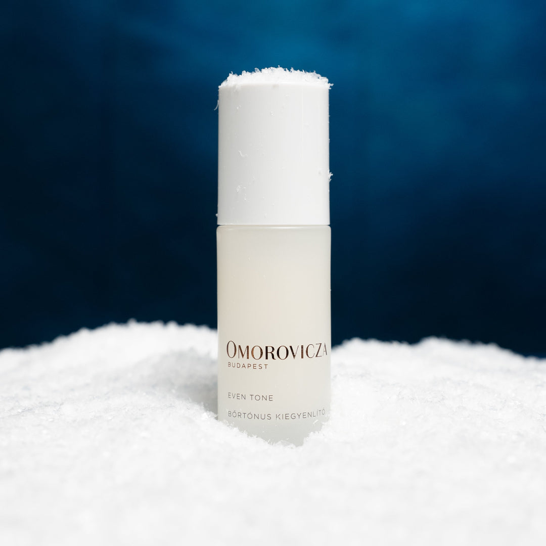 A frosted glass bottle of Omorovicza Even Tone serum with a white cap, standing upright in a bed of white snow against a dark blue background.