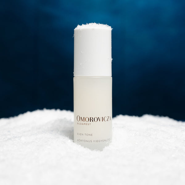 A frosted glass bottle of Omorovicza Even Tone serum with a white cap, standing upright in a bed of white snow against a dark blue background.