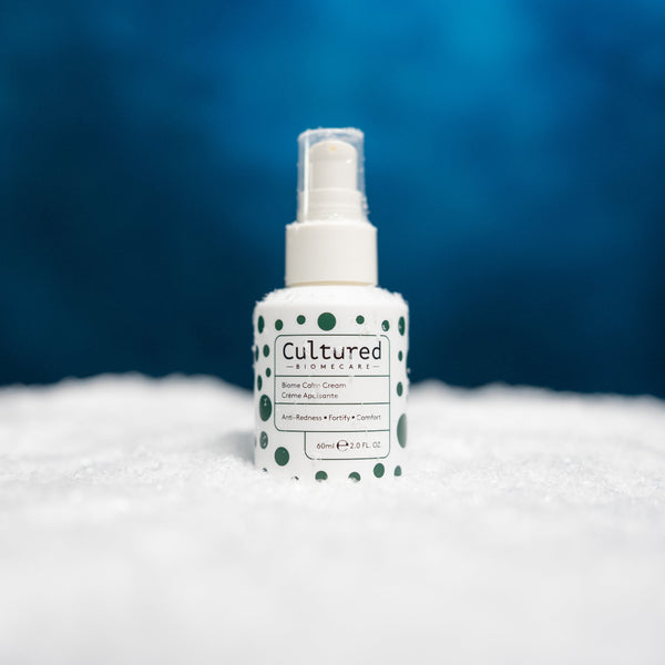 A bottle of Cultured Biomecari Rose Care Cream with green polka dots on a white label, set in a bed of white bath salts against a blue background.